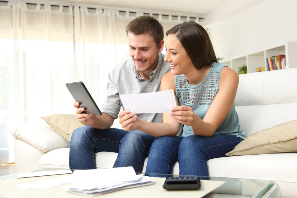 Happy couple checking bank account online in a tablet sitting on a couch in the living room at home